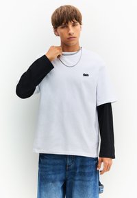 Hvid og sort langærmet t-shirt med logo, featuring et afslappet fit, tekstureret bomuldsstof og lagdelt design. Kombineret med blå jeans.