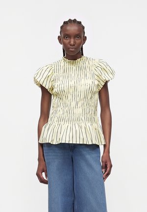 Γυναίκα με σκουρόχρωμο δέρμα και πλεξούδες, που φοράει κίτρινο, ριγωτό τοπ με φουσκωτά μανίκια και φαρδιά μπλε τζιν, στέκεται μπροστά σε λευκό φόντο.