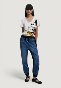 Camiseta blanca de cuello en V con un gran gráfico colorido de Mickey Mouse; combinada con pantalones jogger azules con paneles laterales negros y puños ajustados.