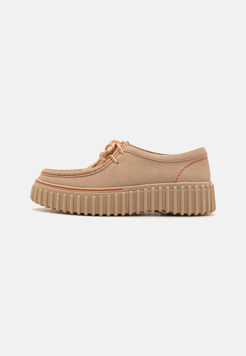 Chaussure en suède beige avec un bout arrondi, des coutures brodées, des détails contrastants orange et une semelle épaisse à nervures.