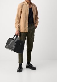 Schwarze Sporttasche in der Hand gehalten, mit glatter Textur, runder Form und doppelt Tragriemen. Getragen mit einem hellbeigen Hemd, grünen Hosen und schwarzen Schuhen.