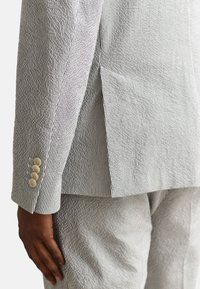 Gros plan sur le torse d’une personne portant un costume rayé gris et blanc texturé avec quatre boutons beiges sur la manche et une fente latérale sur la veste.