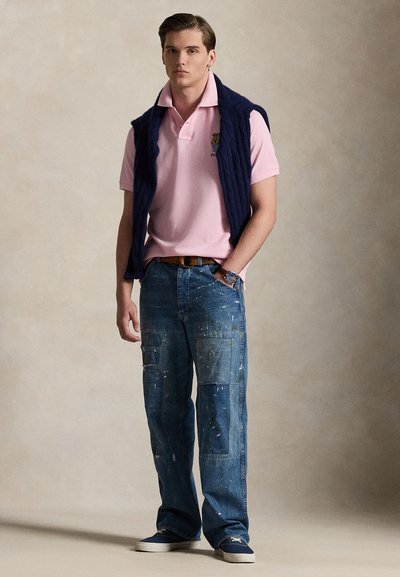 Young man wearing a pink polo shirt, navy sweater draped over shoulders, patched blue jeans, and navy sneakers, standing against beige background.