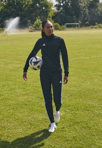 Survêtement noir adidas avec des rayures blanches, un pantalon ajusté et une veste zippée. La personne tient un ballon de soccer noir et blanc.