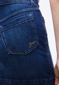 Gros plan sur la poche arrière d'un jean en denim bleu avec le logo "Boss" brodé en or sur la hanche droite, et une main reposant à proximité.