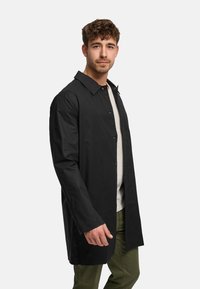 Pitkä musta takki, joka on valmistettu sileästä kankaasta. Takissa on napitettu etuosa, terävä kaulus ja suora leikkaus, ja se on puettu vaalean paidan ja vihreiden housujen päälle.