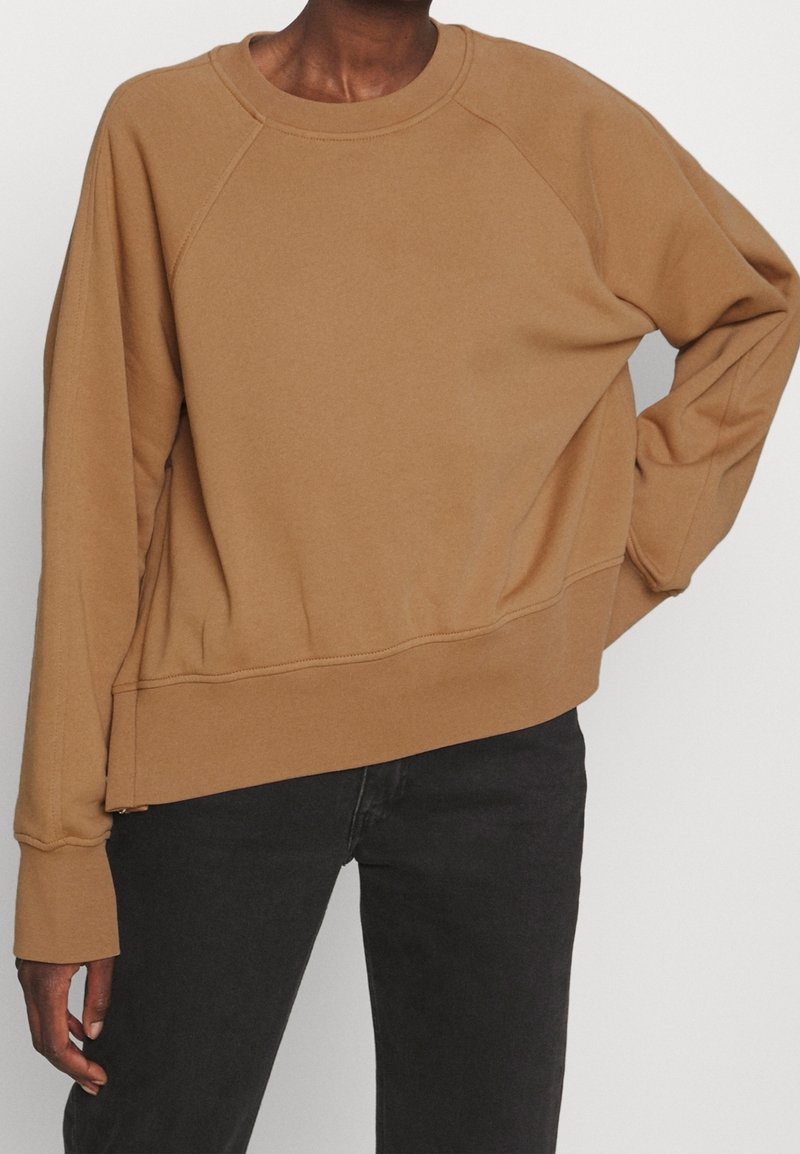 Personne portant un sweat-shirt marron ample avec des manches raglan et un pantalon noir, debout devant un fond clair uni.
