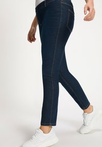 Vaqueros de mezclilla azul oscuro con un corte slim, que presentan una cinturilla elástica, costuras en contraste y dos bolsillos traseros. Combinados con zapatillas blancas.