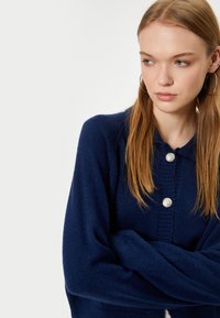 Navyblauer Wollmischungs-Cardigan mit langen Ärmeln, der eine durchgeknöpfte Vorderseite und ein strukturiertes Strickmuster aufweist. Silberne Knöpfe setzen Akzente.