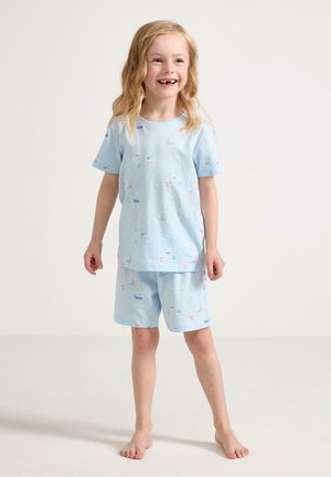 Smilende barfodet barn med langt blond hår iført lyseblå matchende skjorte og shorts med små farverige nautiske tryk.