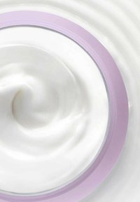 Bocal rond contenant une crème épaisse et blanche avec une texture lisse. Le bocal a un couvercle violet clair avec un bord transparent.