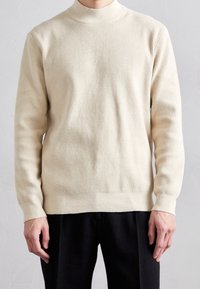 Cremfarbener Strickpullover mit hohem Kragen, geripptem Muster, langen Ärmeln und geradem Saum, kombiniert mit schwarzen Hosen.