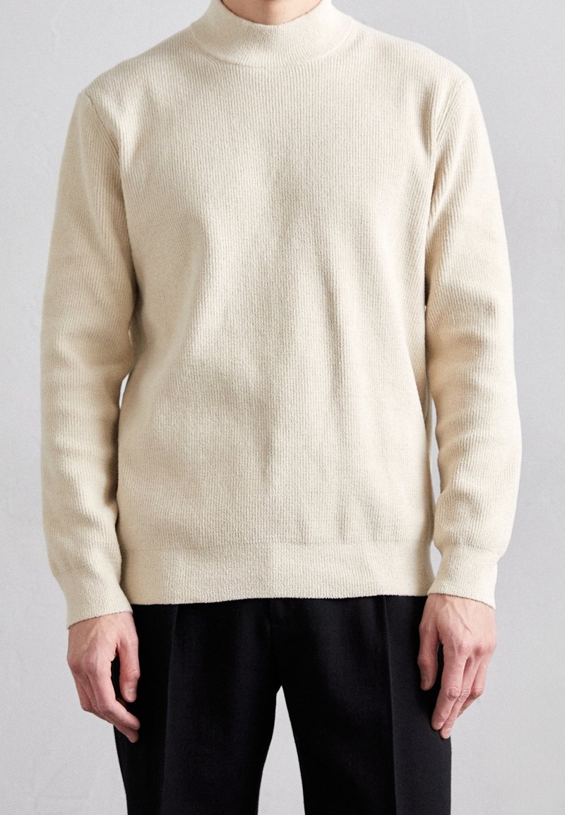 Cremfarbener Strickpullover mit hohem Kragen, geripptem Muster, langen Ärmeln und geradem Saum, kombiniert mit schwarzen Hosen.