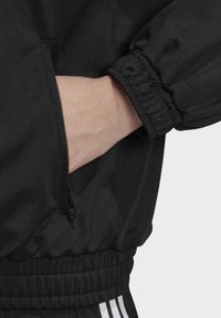 Svart zippered jacka med elastiska ärmslut och midja, med en hand i fickan för att ge en uppfattning om storlek. Slät tyg, minimalisk design.