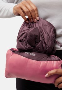 Giacca puffer compatta di un profondo malva, piegata ordinatamente in una morbida busta di tessuto rosa. Presenta un elastico e un'etichetta "STOW AWAY".
