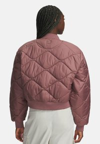 Kobieta z warkoczami, ubrana w mauve'ową pikowaną kurtkę typu bomber i jasne spodnie, pokazana z tyłu na jednolitym tle.