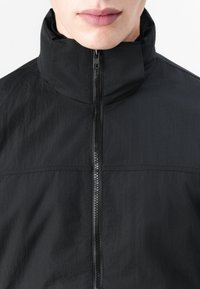 Black zip-up jacket featuring a high collar and a textured fabric with a smooth finish. The zipper closes the front.