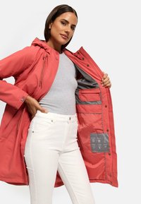 Red waterproof jacket with a hood and drawstring, featuring a gray interior, zippered pockets, and care label details. Paired with white jeans.