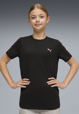 Młoda osoba z rękami na biodrach, ubrana w czarną koszulkę Puma z różowym logo i paskami na rękawach, szare tło.