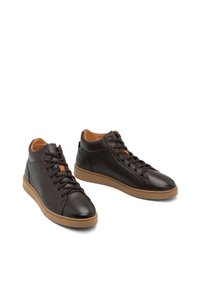 Un par de zapatillas altas de cuero marrón oscuro con suelas de goma beige y cordones marrones sobre un fondo blanco.