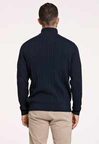 Marineblauwe kabelgebreide trui met een hoge kraag, geribde manchetten en een lichte textuur. Eenvoudig ontwerp met een standaard pasvorm en geen zichtbare logo's.