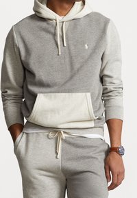 Grå hoodie i bomullsmix med en krämfärgad huva, känguruficka och dragsko. Har en liten logotyp på bröstet och mjuka texturer.