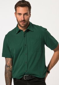 Camisa de botones de manga corta en tejido verde oscuro. Presenta un cuello, un bolsillo delantero y botones negros. Modelo lleva pantalones negros.