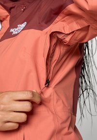 Lett koralljakke med maroon øvre del, som har en glidelåslomme med en trekksnor. Stoffet har en glatt tekstur.