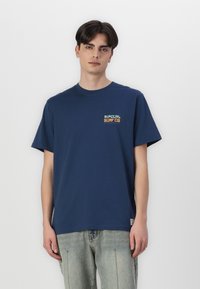Mørkeblå bomulds t-shirt med et lille "Rip Curl Surf Co" logo i orange og grønt på venstre bryst, parret med lyse denim jeans.