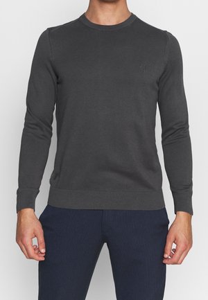 Man draagt een aangesloten donkergrijze trui met lange mouwen en ronde hals, en marineblauwe broek met krijtstrepen, staand met ontspannen armen.
