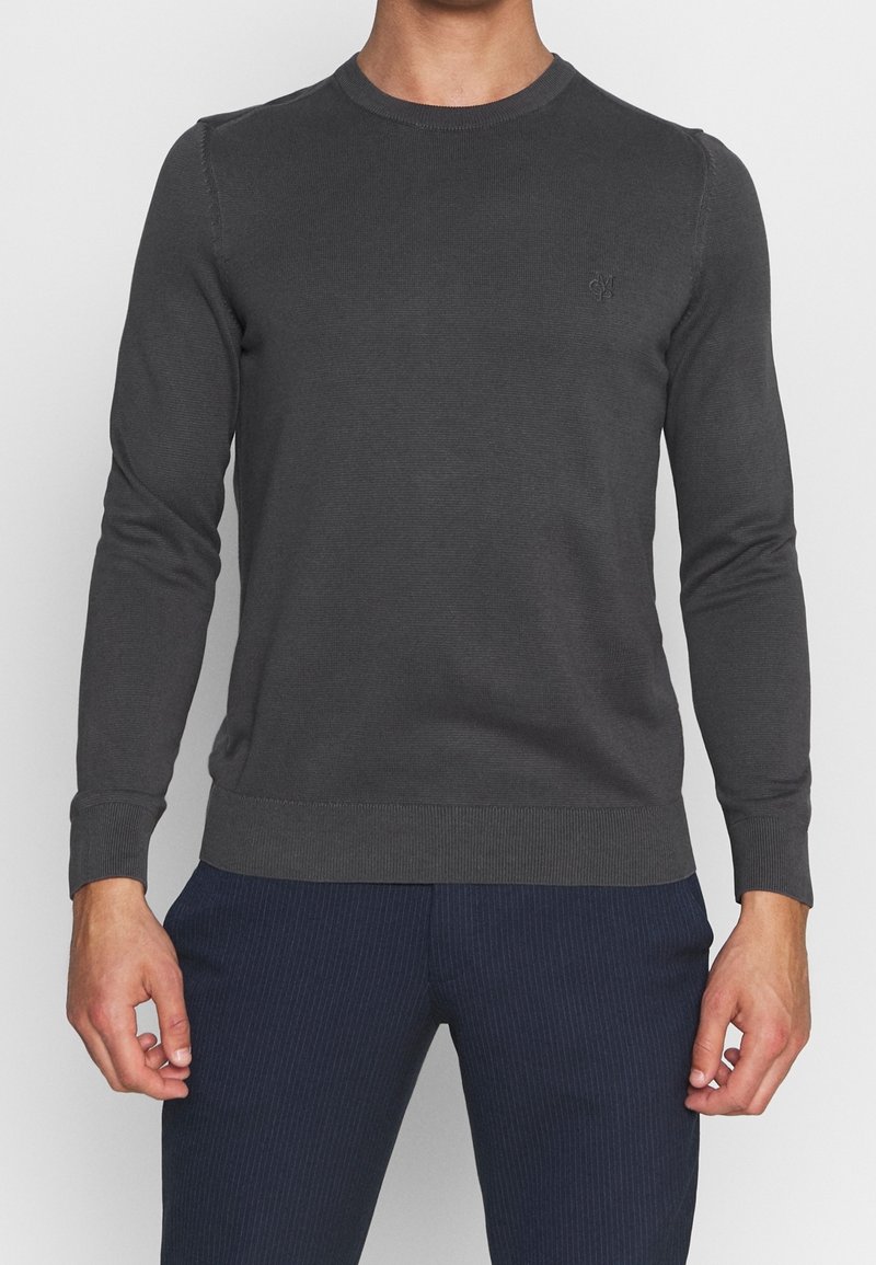 Homme portant un pull gris foncé ajusté à manches longues avec col rond et un pantalon bleu marine à fines rayures, debout, les bras relâchés.
