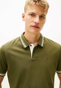 oliwkowa koszulka polo z teksturowanej tkaniny, białym kołnierzykiem i akcentami w paski. Zawiera małe logo na klatce piersiowej.