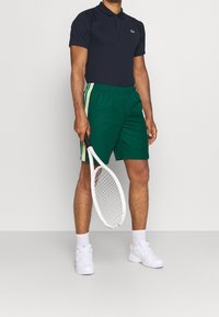 Marineblaues Poloshirt mit kurzen Ärmeln, grüne Shorts mit weißen und gelben Seitenstreifen und weiße Sneakers, hält einen weißen Tennisschläger.