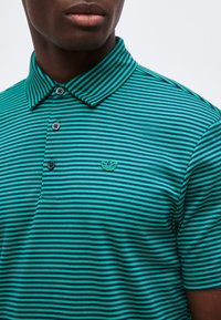 Κοντινό πλάνο ενός ατόμου που φοράει πράσινη και σκούρο μπλε ριγέ πόλο μπλούζα με τρία κουμπιά και μικρό κεντητό λογότυπο στο στήθος.