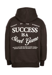 Hoodie marron foncé avec un texte blanc au dos indiquant : « Enseigné par les rues. Le succès est un jeu de rue. Rêve grand, travaille dur et fais en sorte que cela arrive. »