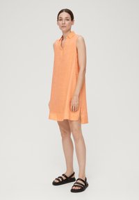 Ärmelloses Kleid aus weichem orangefarbenem Leinen mit einem Kragen, seitlichen Schlitzen und einer lockeren Passform, kombiniert mit schwarzen Riemchensandalen.