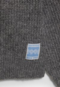 Sivi pleteni pulover s teksturiranom tkaninom. Ima plavu oznaku s natpisom "KNITTERS 99" i "SINCE 1991" u bijelom fontu.