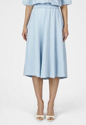 Frau trägt einen hellblauen Midirock mit elastischem Bund und weiße offene High Heels, steht vor einem einfarbigen Hintergrund.