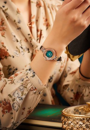 Reloj de pulsera redondo con una esfera color teal, acentos en tono plateado y una correa de cuero negra. Las características incluyen un bisel estampado y pequeñas piedras decorativas.