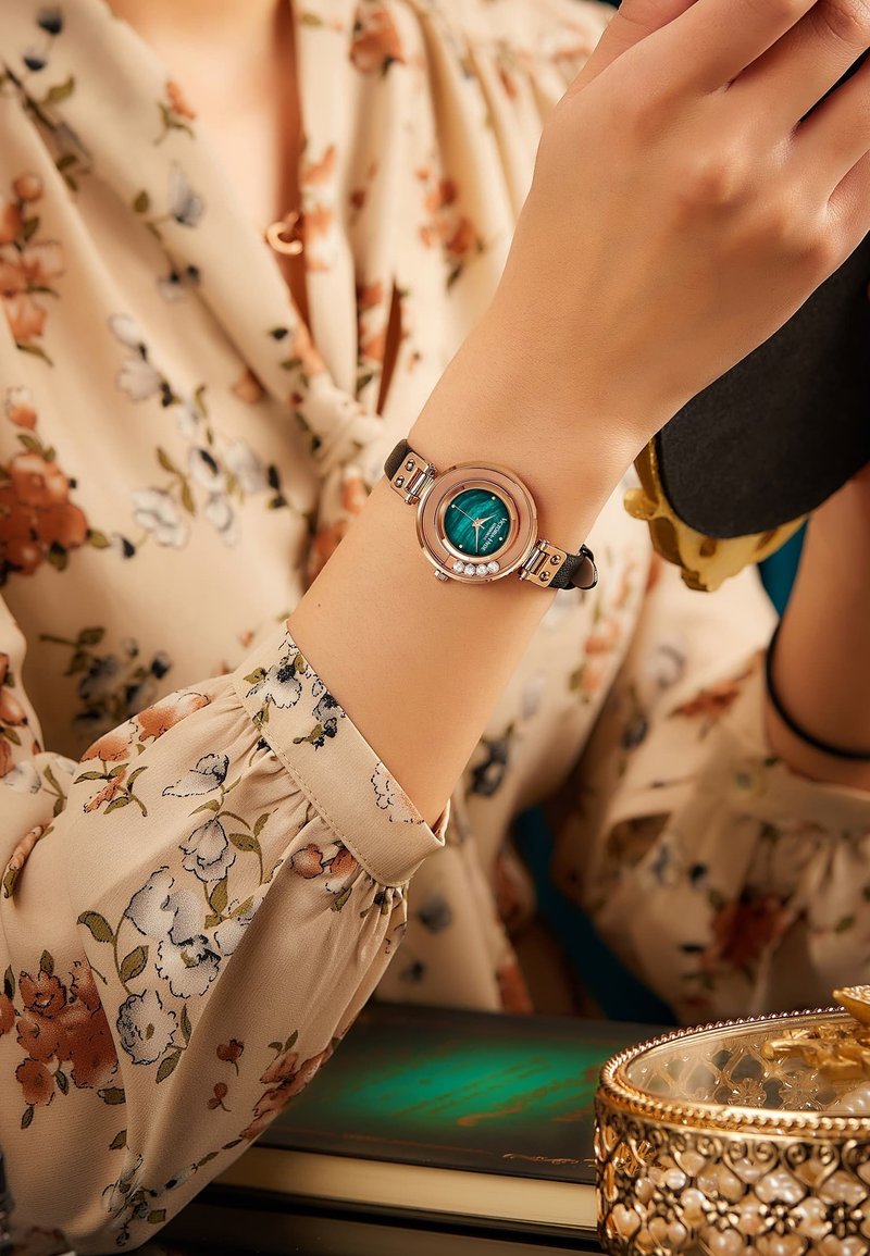 Reloj de pulsera redondo con una esfera color teal, acentos en tono plateado y una correa de cuero negra. Las características incluyen un bisel estampado y pequeñas piedras decorativas.