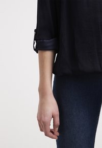 Svart, lätt, långärmad skjorta med uppkavlade manschetter, ihop med blå denimjeans. Fokusera på handen som vilar vid tyget.