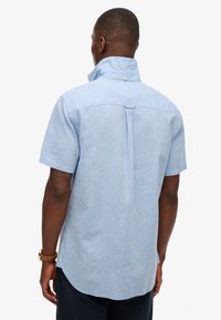 Koszula z krótkim rękawem w jasnoniebieskim kolorze, z kołnierzykiem stójkowym, detalem w postaci fałdy z tyłu i zaokrąglonym dołem. Materiał wydaje się lekki i lekko strukturalny.