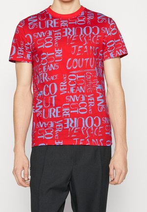 Person iført en rød t-shirt med korte ærmer med spredt blå tekst, der læser "Versace Jeans Couture", og sorte bukser.