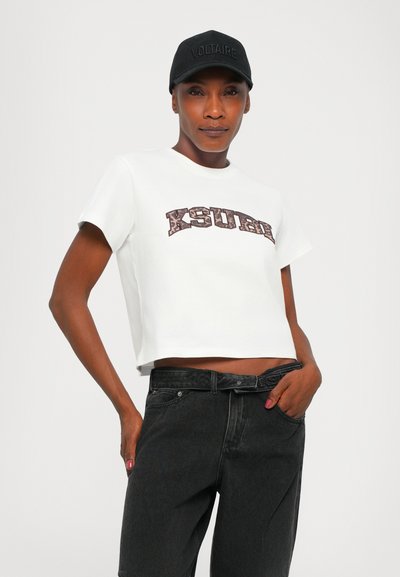 Camiseta blanca corta con el texto "KSURE" en un patrón marrón. Combinada con jeans negros. Sombrero negro con la inscripción "VOLTAIRE." Textura de tela suave.