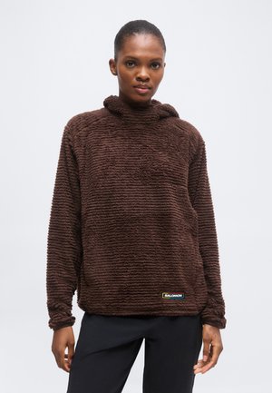 Frau trägt einen dunkelbraunen, strukturierten Fleece-Pullover mit kleinem Salomon-Logo nahe dem unteren Rand, steht vor einem einfarbigen Hintergrund.