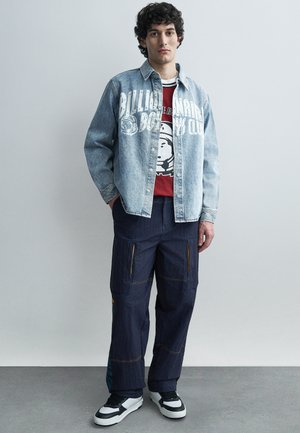 Fiatal férfi áll, világos farmerdzsekit visel nagy fehér felirattal, piros grafikás pólót, sötét cargo nadrágot és fekete-fehér tornacipőt.