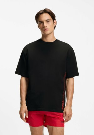 Jeune homme portant un T-shirt noir ample avec un texte rouge "HUGO" sur le côté et un short rouge sur fond blanc.