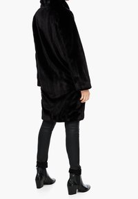 Černý kabát z umělé kožešiny s vysokým límcem, dlouhými rukávy a volným střihem. V kombinaci s černými koženými kalhotami a botami na podpatku s texturou po kotníky.