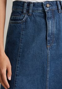 Jupe en denim bleu foncé avec une silhouette droite. Présente une fermeture par boutons à l'avant, des coutures surpiquées et une poche latérale.