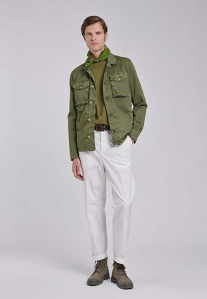 Man draagt een olijfgroene jas, bijpassende sjaal, olijfgroen hemd, witte broek en bruine laarzen, staand met één hand in de zak tegen een effen achtergrond.